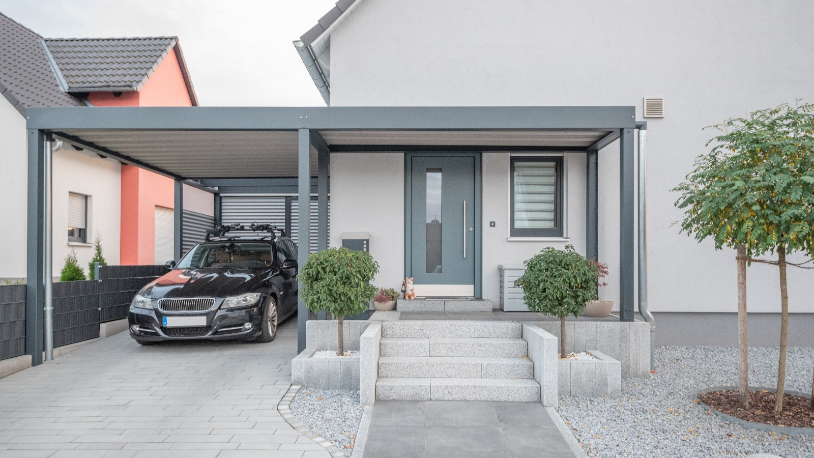 Modern gestalteter Carport von BESTBAU, bietet stilvollen und effektiven Schutz für Autos, integriert in das Design eines Einfamilienhauses mit gepflegter Einfahrt und Zierpflanzen.